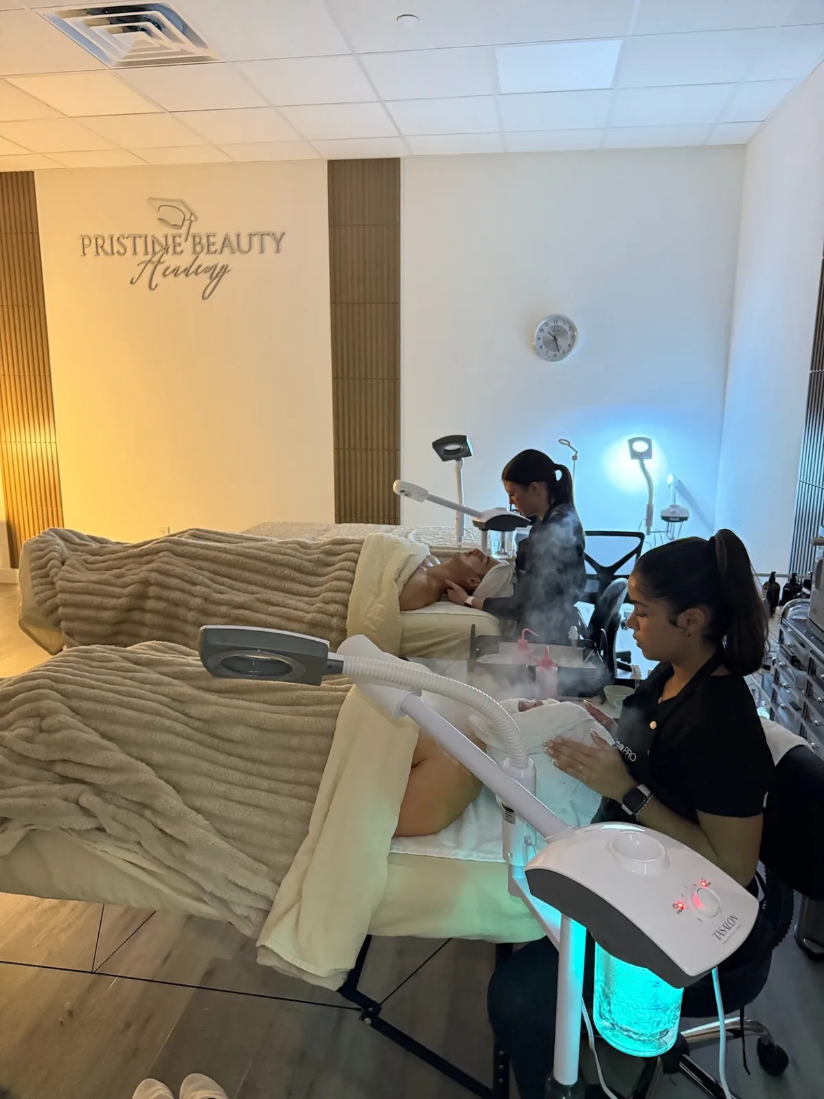 Two students working on clients in the two-bed treatment room under the Pristine Beauty Academy sign
